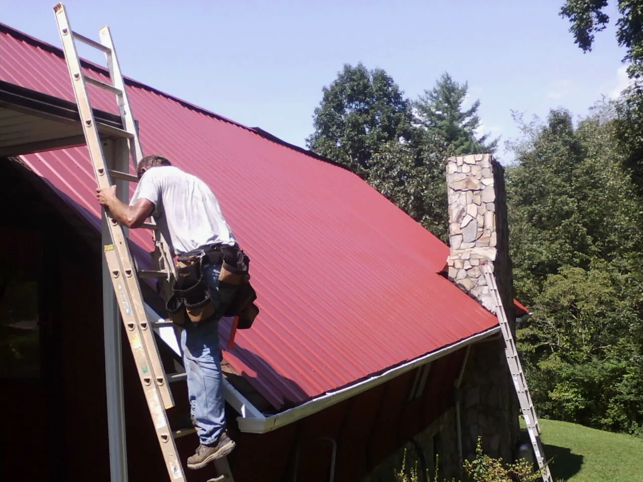 Roofer on a ladder beside a red metal roof and stone chimney — Soffit & Fascia Repair by Roofing Brothers in Maineville